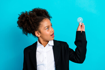 Young schoolgirl in formal outfit holding light bulb against teal background, representing creativity and school education