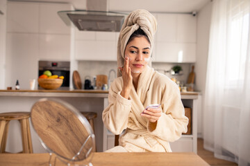 Happy Woman in Beige Robe and Towel Caring for Skin in Bright Modern Kitchen