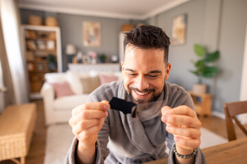 Obraz premium Happy Bearded Man In Grey Sweater Examining Small Item With Excitement In Modern Home Interior