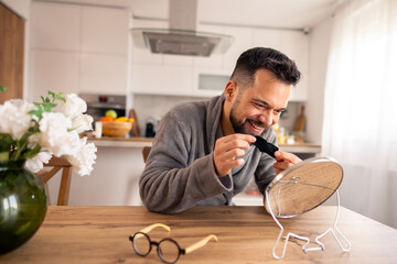 Smiling Caucasian Man in Grey Robe Applies Cream to Face in Brightly Lit Kitchen