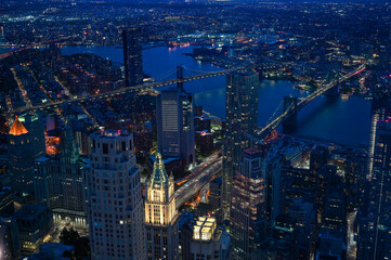 Aerial night view of Lower Manhattan in New York City with illuminated skyscrapers, the Brooklyn Bridge and East River, city lights stretching into the distance.