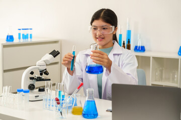 Fototapeta premium Asian schoolgirl wearing lab coat and goggles smiles while holding beaker and test tube in science classroom, reflecting joy and excitement in hands-on laboratory learning experience.