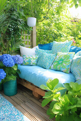 Vertical Photo of Boho Outdoor Sofa on Garden Terrace, Surrounded by Lush Greenery and Blue Hydrangea Flowers, Cozy Patio or Balcony Summer Decor, Natural Relaxation and Stylish Backyard Inspiration.