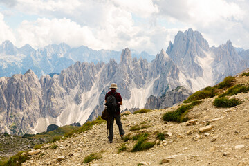 Fototapeta premium Rear view of male tourist with knapsack on shoulders and trekking pole in hands standing on mountain slope, enjoying scenic view of rugged rocky peaks of Dolomites under cloudy sky