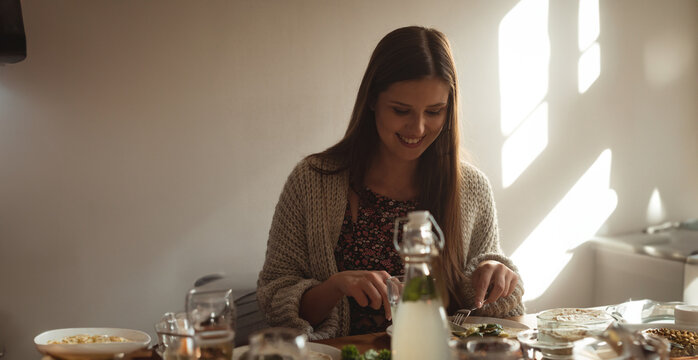 Woman cutting salad and preparing meal on wooden dining table near glass bottle and dishes