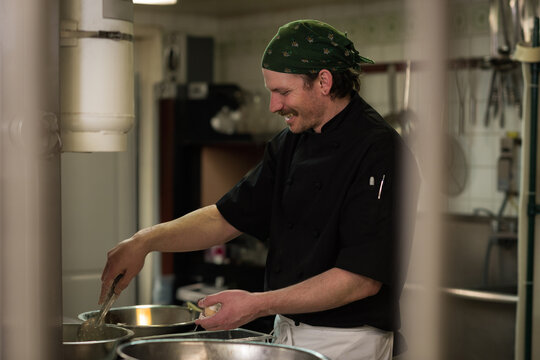 Mid-adult chef wearing chef coat green bandana ladling food into bowls at prep station in kitchen