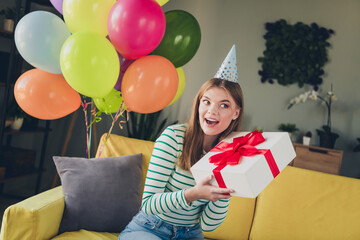 Cheerful young woman celebrating indoors with balloons and a gift, wearing a party hat and striped...