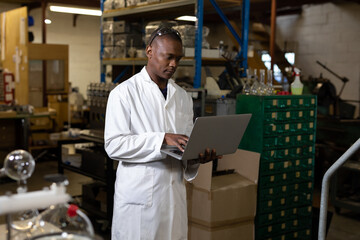 African American man wearing white lab coat typing on laptop in lab workshop near metal shelving