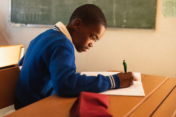 African American schoolboy leaning over writing in open notebook with green pen on classroom desk