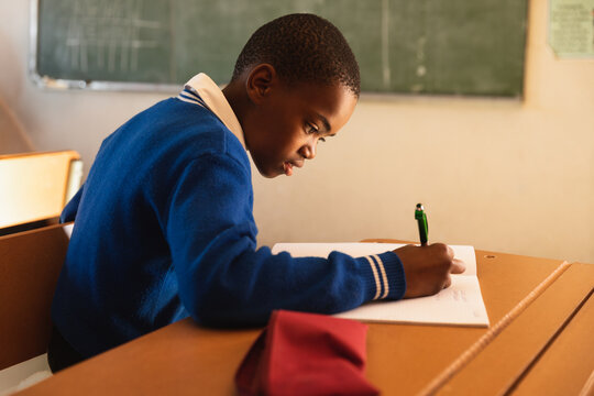 African American schoolboy leaning over writing in open notebook with green pen on classroom desk