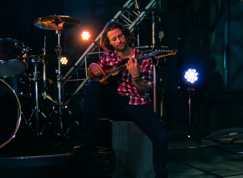 Man in his thirties sitting on speaker playing electric guitar on club stage under colored lights