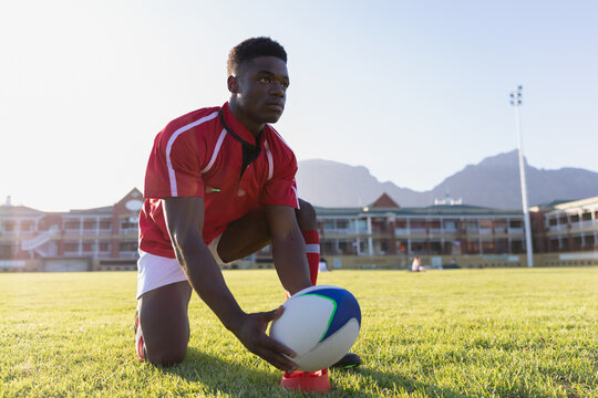 African American male athlete kneeling on pitch near clubhouse positioning rugby ball on red tee