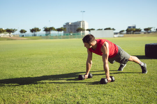 Athletic man performing push-ups using hexagonal dumbbells on grassy sports field near tractor tire