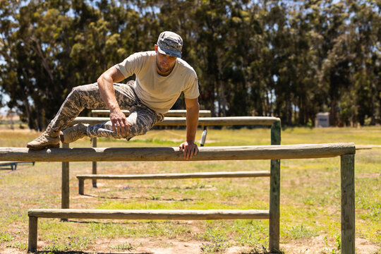 Male soldier in camouflage pants and tan t-shirt vaulting over wooden hurdle on training ground