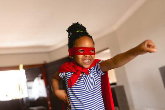 African American child girl flying in home living room wearing red superhero mask and cape