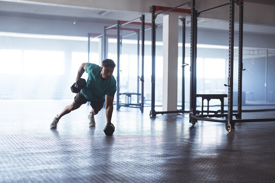 Man doing plank row holding hex dumbbell on rubber mat near squat rack and pull-up rig