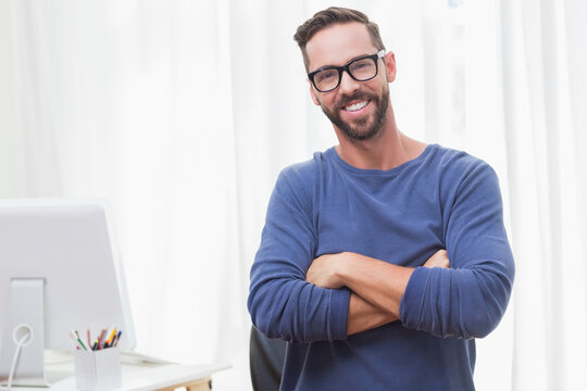 Man in blue shirt standing at office desk holding colored pencil cup, looking at computer monitor