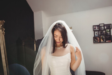 Asian bride standing in bridal dressing room holding tulle veil at shoulder wearing wedding gown