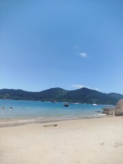 Fototapeta premium View of a sandy Ubatuba beach with mountains in the background and boats on the water under a clear blue sky