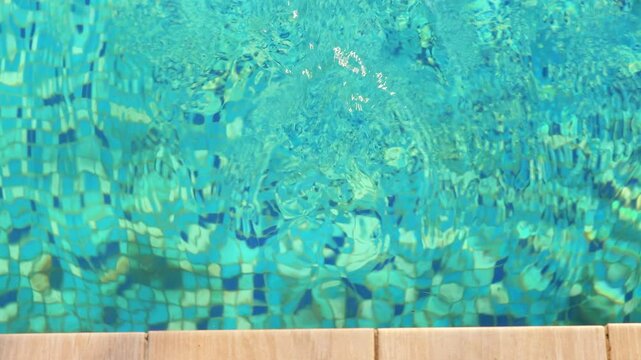 An overhead view of a vibrant blue swimming pool with shimmering water and visible mosaic tiles at the bottom, bordered by light wood decking.