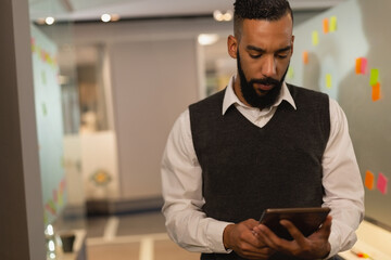 African American man in sweater vest holding tablet reviewing sticky notes at workspace, copy space
