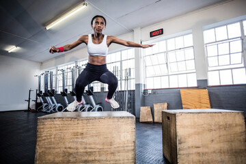 African American woman jumping onto wooden plyometric box under digital clock in CrossFit studio