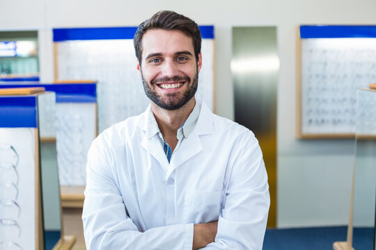 Male optician wearing lab coat standing in optical showroom, smiling near eyewear displays