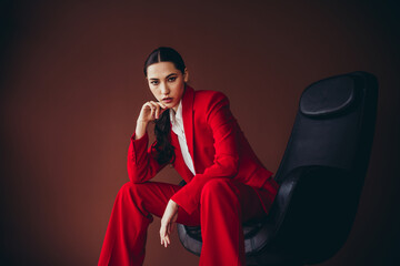 Attractive Elegant Woman in Red Business Suit Seated Confidently Against a Brown Background