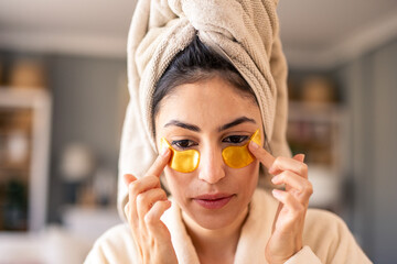 Happy Woman in Beige Robe with Towel on Head Applying Gold Eye Patches in Gentle Lighting