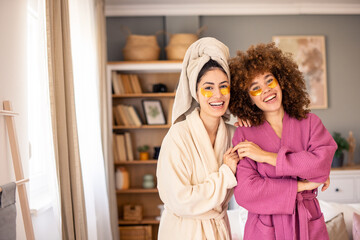 Two Smiling Women in Bathrobes with Bright Under-Eye Patches in a Warm, Relaxing Room