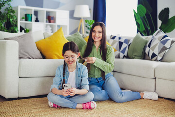 Young woman braiding her daughter's hair while sitting together on the floor in their comfortable and cozy living room