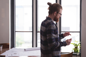 Transparent tablet resting on desk beside rolled blueprints and steaming coffee cup near window