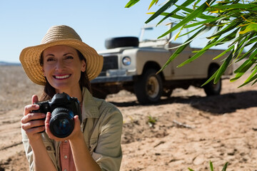 Woman photographer posing in sandy field holding DSLR camera under straw hat beside SUV, copy space