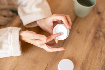 Smiling Woman in Beige Robe Applying Skincare Cream with Wooden Table and Green Cup in Background