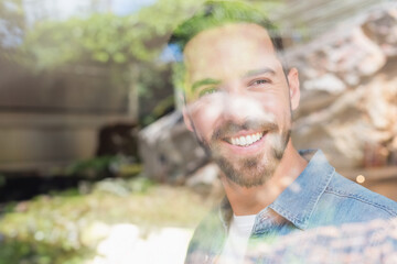 Mid adult male wearing white t-shirt and denim shirt standing inside home behind glass pane smiling