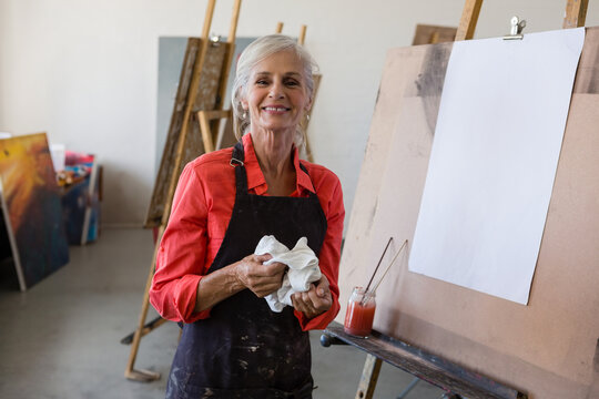 Senior female artist wearing red shirt apron smiling and holding cloth in studio with easels