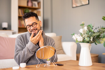 Happy Asian Man With Glasses Applying Face Cream During Morning Self Care Routine In Modern Living Room Setting