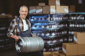 Smiling mature adult man carrying metal keg in warehouse with shelving, bottled cases and boxes