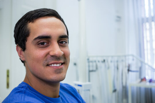 Man in blue scrubs smiling in staff room with coat rack and door handle, copy space