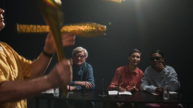 Rack focus of diverse members of jury sitting at table with microphones and discussing performance of skilled mature Caucasian male juggler tossing clubs auditioning for talent show