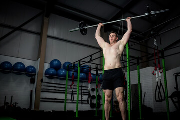 Shirtless man lifting barbell overhead in gym by squat rack, stability balls, resistance bands