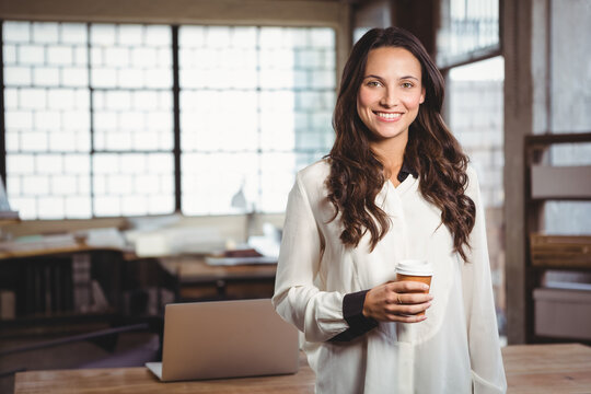 Adult woman holding paper coffee cup wearing blouse standing beside open laptop on desk in office