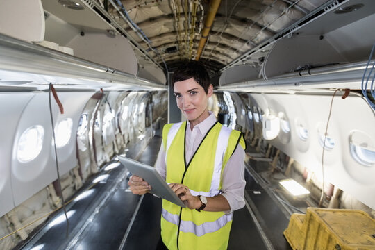 Middle-aged woman wearing high-vis vest inspecting wiring and holding tablet in aircraft cabin