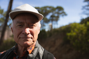 Senior man standing on wooded trail wearing baseball cap and quilted vest with backpack