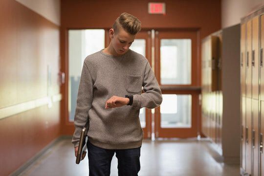 Chinese teenage male student walking by lockers in high school checking smartwatch, carrying laptop