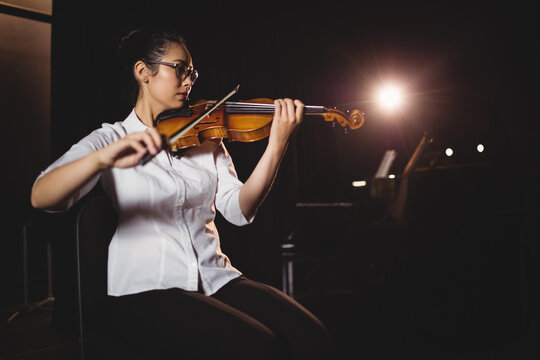 Violin being played on dimly lit stage under warm spotlight, with grand piano in shadows