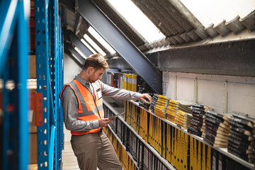 Standing in warehouse aisle male wearing safety vest reaching for binder with tablet, copy space