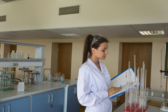 Female chemist wearing white lab coat consulting clipboard at laboratory bench with glassware