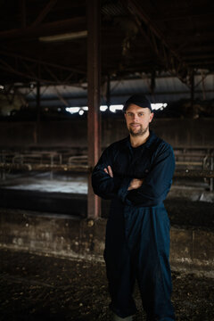 Male farm caretaker wearing coveralls standing with arms crossed in barn among metal feeding stalls