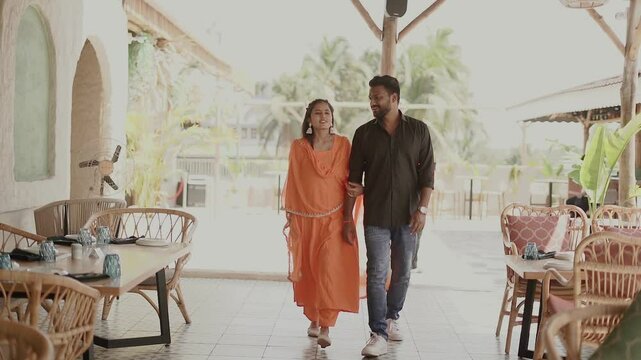An affectionate Indian couple shares an intimate moment outdoors, dressed in traditional ethnic attire. The woman wears a bright orange salwar suit while the man is in a black shirt and jeans. 